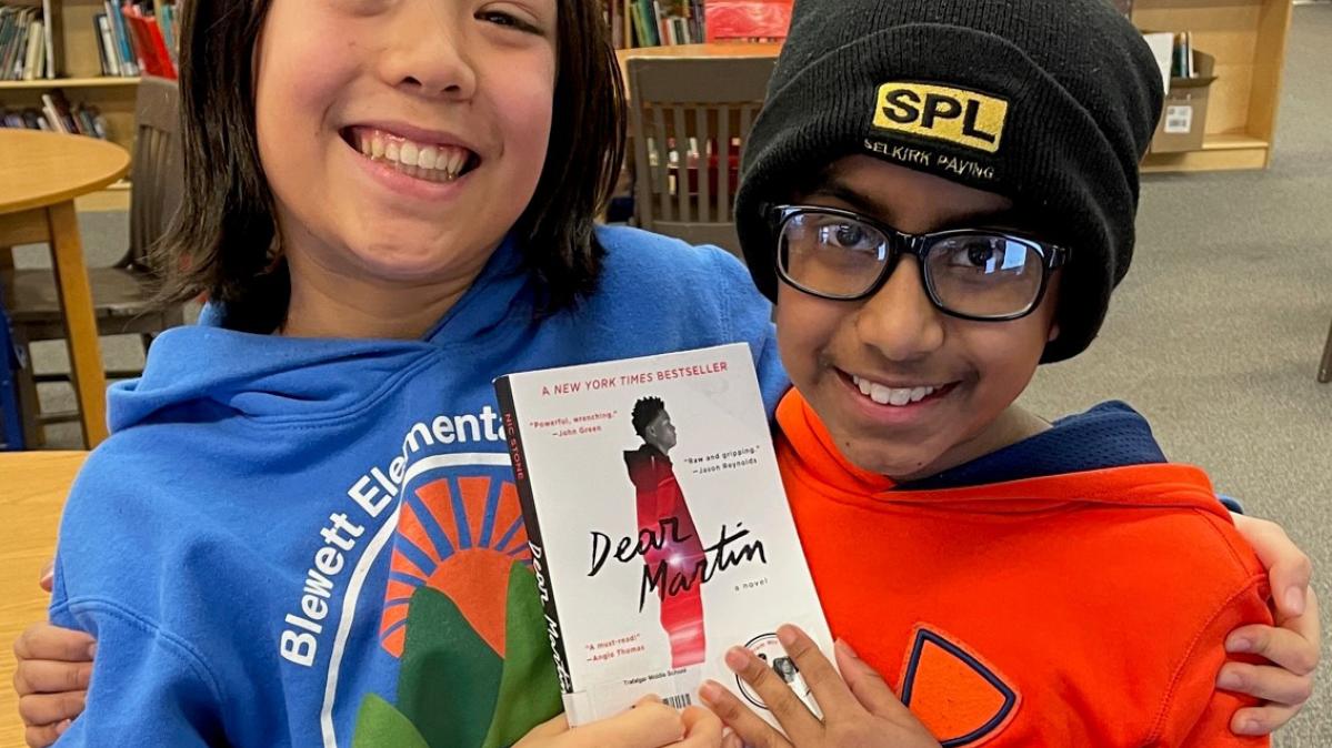 One boy in a blue sweatshirt and one in a red sweatshirt are both smiling widely with arms around one another's shoulders, holding a book up together in a library.