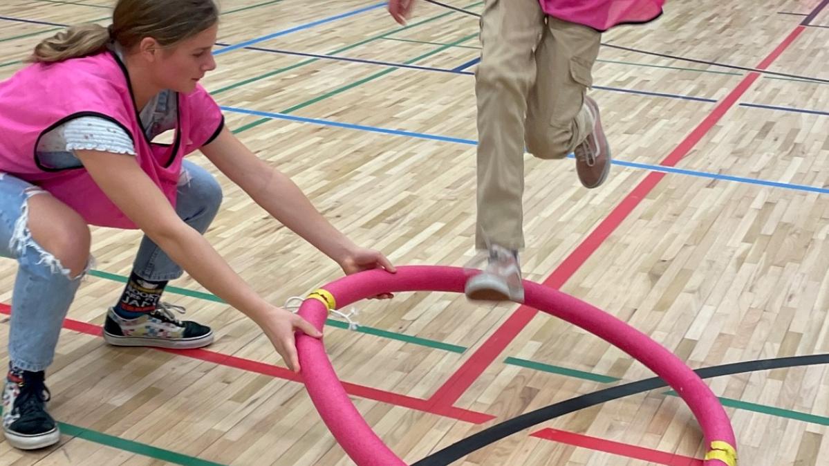 Two girls in pink tunics participate in an indoor game in a school gymnasium with one girl holding a pink hoola hoop on the ground and the other leaping into it. Other students look on.