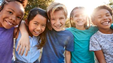 Five diverse, smiling intermediate grade students arm in arm against the backdrop of a sunrise through leafy green trees 